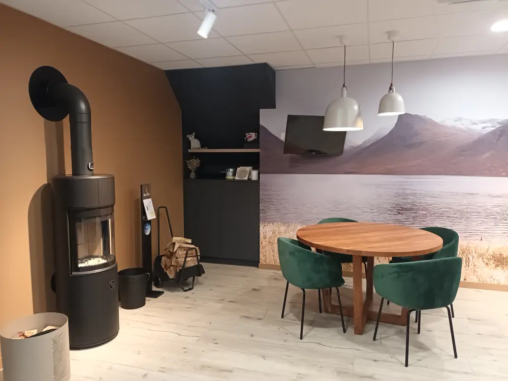 Photo présentant l'espace rencontre du showroom, avec un poêle à bûches près d'une table ronde en bois entourée de 4 fauteuils verts en velours.